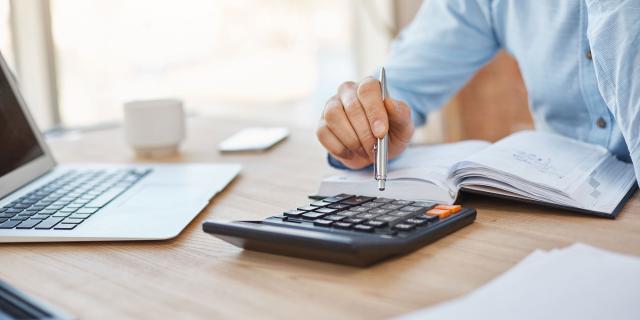 Close up detail of professional serious accountant sitting in light office, checking company finance profits on calculator, writing down results in notebook. Business concept.