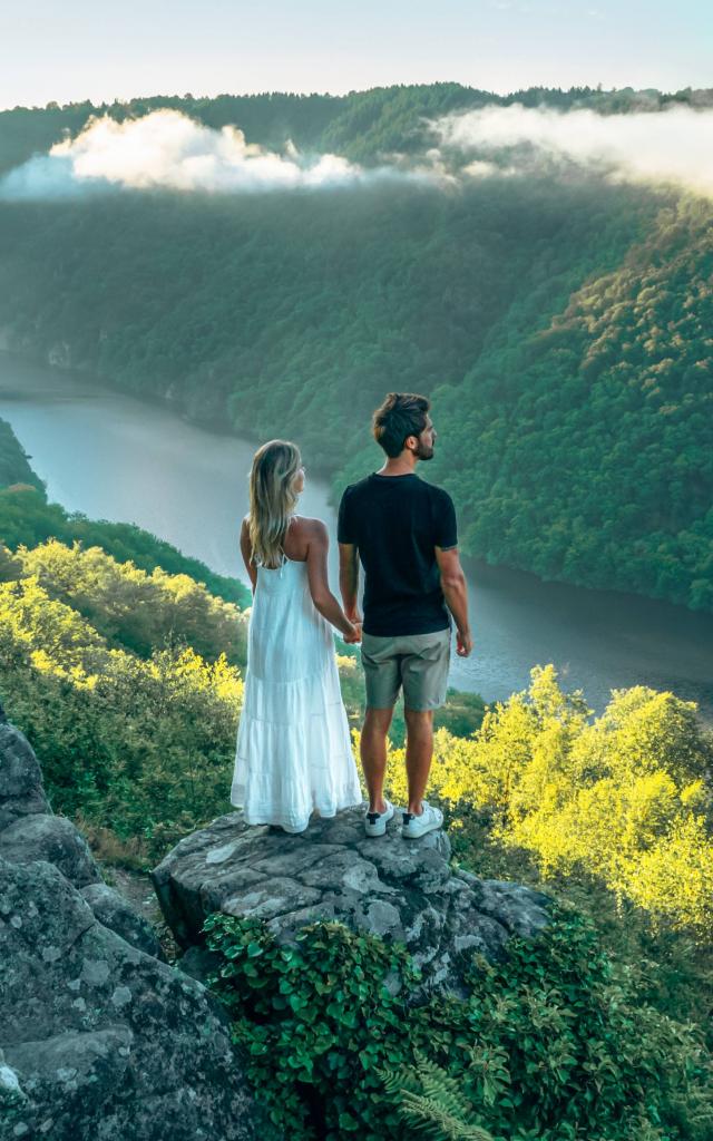 Une femme blonde avec une robe blanche et un homme brun en tee-shirt noir et short gris, se tiennent la main et regardent la rivière Dordogne et ses gorges depuis le point de vue du Roc du Busatier.