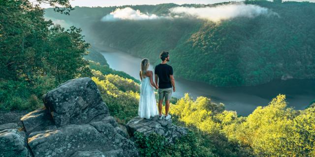 Une femme blonde avec une robe blanche et un homme brun en tee-shirt noir et short gris, se tiennent la main et regardent la rivière Dordogne et ses gorges depuis le point de vue du Roc du Busatier.