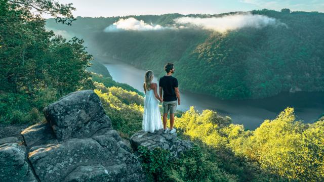 Une femme blonde avec une robe blanche et un homme brun en tee-shirt noir et short gris, se tiennent la main et regardent la rivière Dordogne et ses gorges depuis le point de vue du Roc du Busatier.