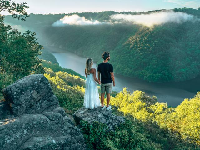 Une femme blonde avec une robe blanche et un homme brun en tee-shirt noir et short gris, se tiennent la main et regardent la rivière Dordogne et ses gorges depuis le point de vue du Roc du Busatier.