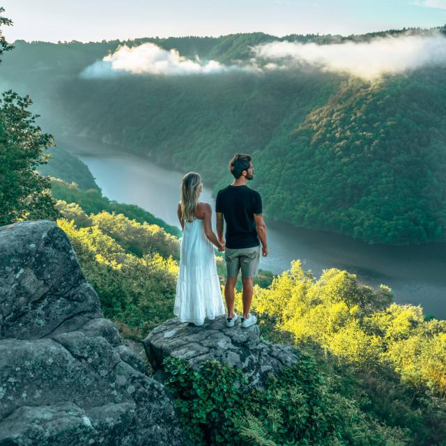 Une femme blonde avec une robe blanche et un homme brun en tee-shirt noir et short gris, se tiennent la main et regardent la rivière Dordogne et ses gorges depuis le point de vue du Roc du Busatier.